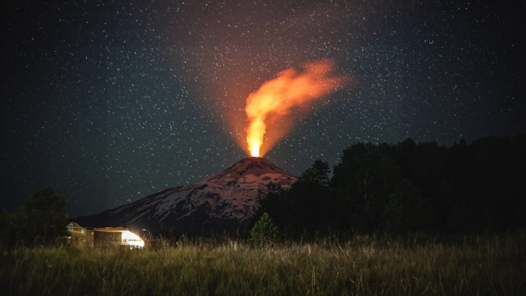 Ein nächtlicher Vulkan bricht aus, glühend orangefarbene Lava und Rauch steigen in einen sternenübersäten Himmel. Der Berg ist mit Schnee bedeckt, und im grasbewachsenen Vordergrund leuchtet ein kleines Gebäude.