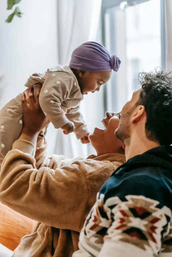 Ein fröhliches Paar lächelt und hebt ein Baby in einem gepunkteten Strampler und einem lilafarbenen Kopftuch in der Nähe eines sonnenbeschienenen Fensters hoch, um einen glücklichen, spielerischen Moment miteinander zu verbringen.