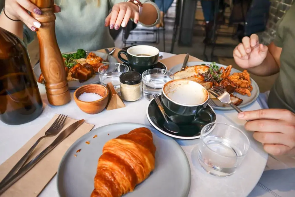 Zwei Personen sitzen an einem Tisch mit Tellern voller Essen, darunter ein Croissant, Salat und gebratene Sachen. Tassen mit Kaffee, Gläser mit Wasser und Gewürze stehen ebenfalls auf dem Tisch. Eine Person benutzt eine Pfeffermühle.
