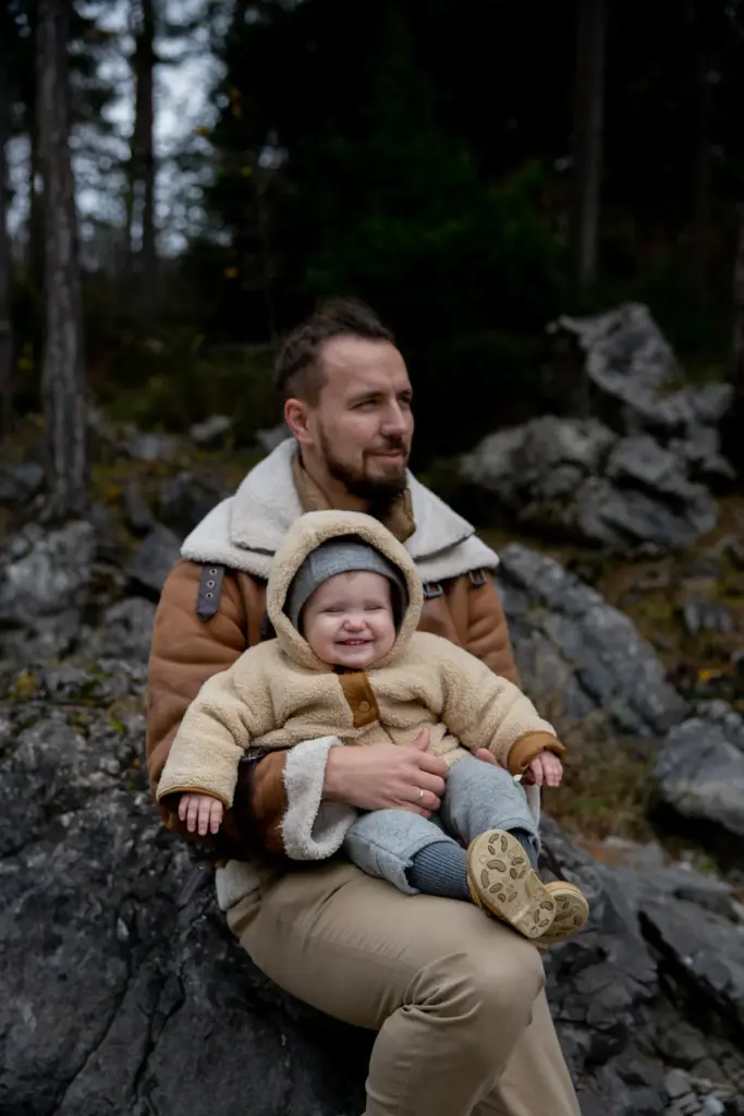 Ein Mann in einer braunen Jacke sitzt auf Felsen in einem Wald und hält ein lächelndes Baby in einem hellbraunen Mantel, einer grauen Hose und einem grauen Hut. Beide sind warm gekleidet und scheinen zufrieden im Freien zu sitzen.