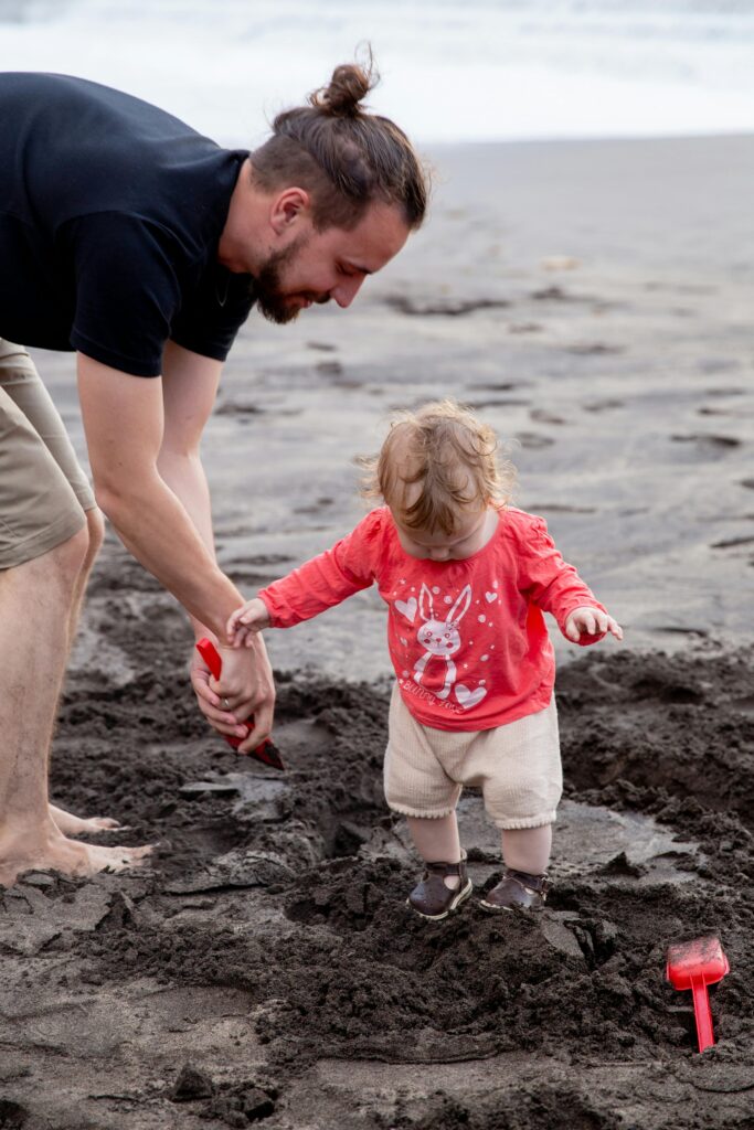 Ein Mann hilft einem kleinen Kind, an einem Sandstrand zu stehen. Das Kind trägt ein rotes Hemd mit einer Hasengrafik und helle Shorts, während der Mann barfuß ist und eine rote Schaufel hält. Eine rote Spielzeugschaufel liegt in der Nähe im Sand.