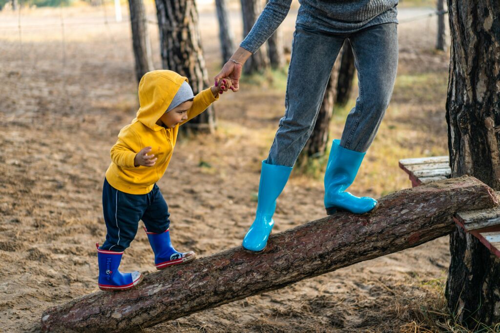 Ein Kind in einem gelben Kapuzenpulli und blauen Stiefeln läuft auf einem umgestürzten Baumstamm und hält sich an der Hand eines Erwachsenen fest. Der Erwachsene trägt blaue Stiefel und graue Kleidung. Die Szene spielt sich im Freien zwischen Bäumen ab.