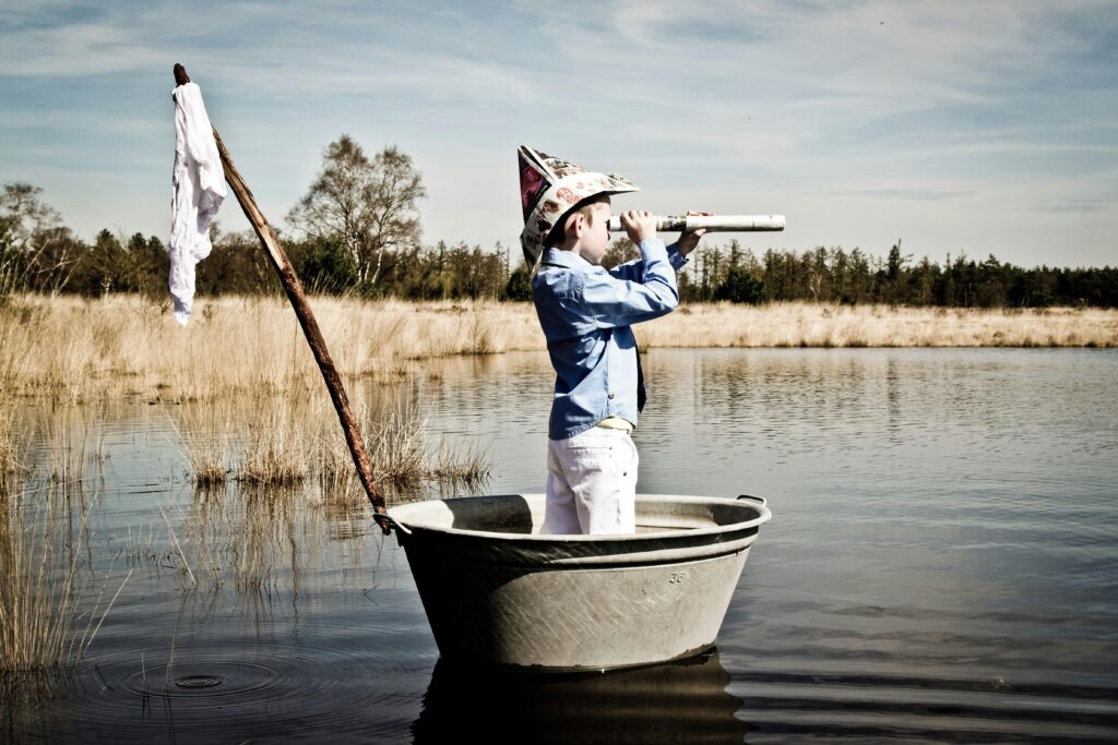 Eine Person mit einem Papierhut steht in einem kleinen Metallboot auf einem See und schaut durch ein Fernrohr. Das Boot hat eine provisorische Flagge, und die Szene ist von hohen Gräsern und Bäumen im Hintergrund umgeben.