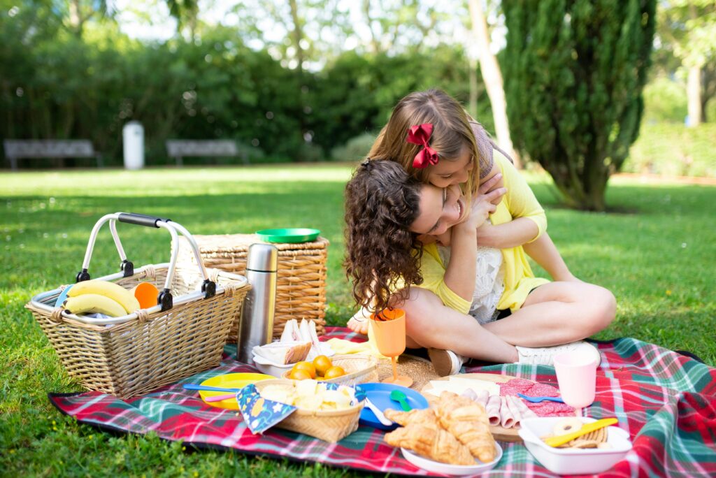 Zwei Kinder sitzen auf einer Picknickdecke in einem Park, umarmen sich und lächeln. Picknick-Essen, Teller, Bananen und Körbe sind um sie herum auf der Decke ausgebreitet. Im Hintergrund sind grünes Gras und Bäume zu sehen.