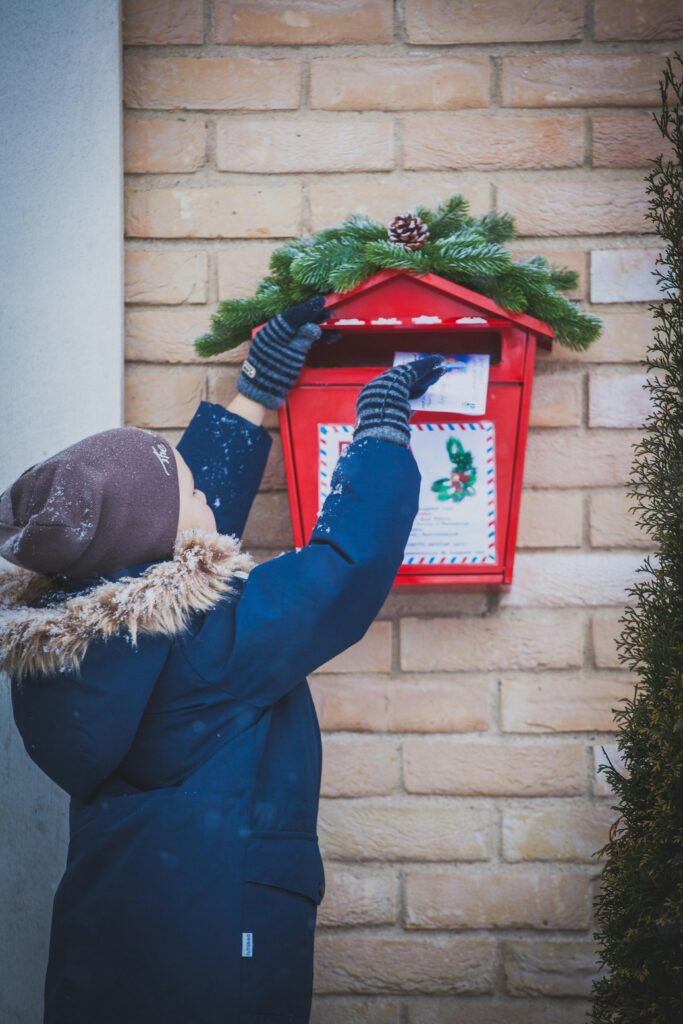 Eine Person in Wintermantel und Strickmütze wirft einen Brief in einen festlich geschmückten roten Briefkasten, der mit Grünzeug und einem Tannenzapfen verziert ist und an einer Backsteinmauer angebracht ist. Auf der Kleidung der Person ist Schnee zu sehen.