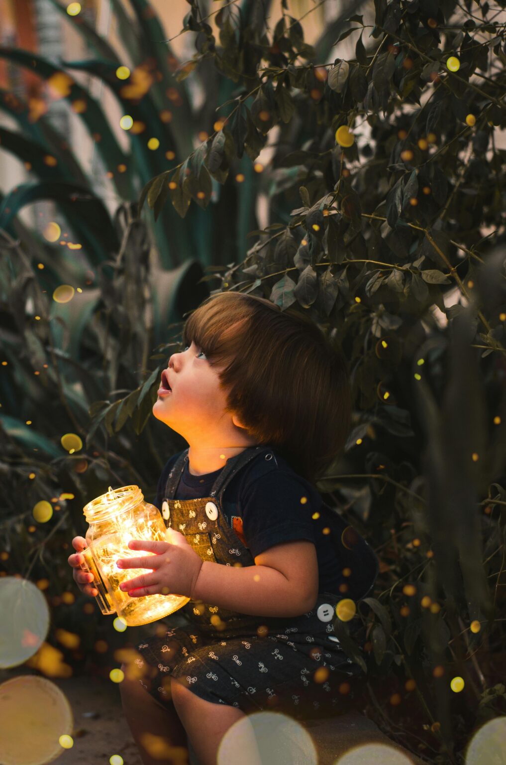 Ein kleines Kind mit braunen Haaren sitzt in der Abenddämmerung im Freien zwischen Pflanzen und blickt staunend nach oben, während es ein leuchtendes Glas mit warmen goldenen Lichtern in der Hand hält, umgeben von weichen, magischen gelben Lichtkugeln.