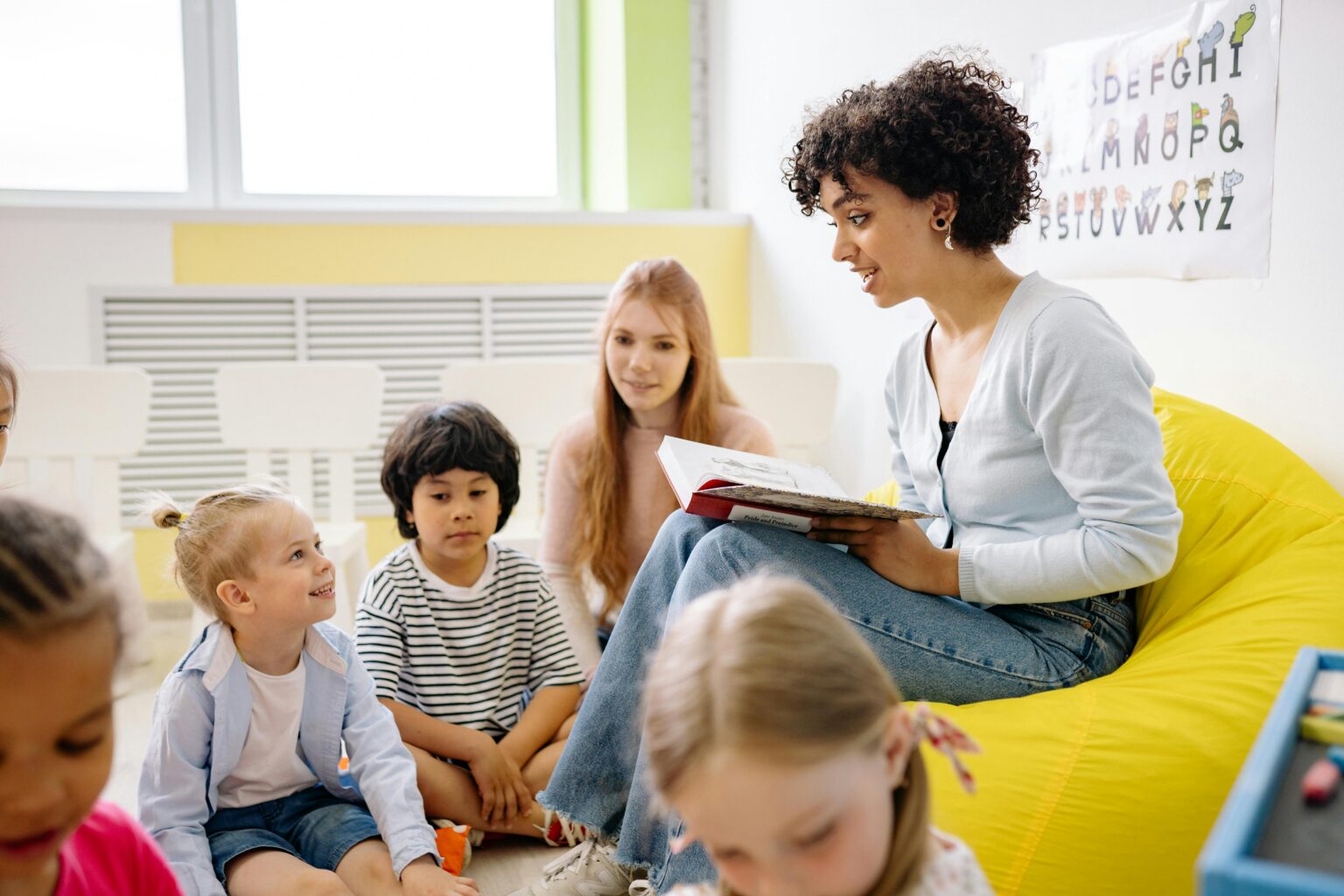 Eine Frau liest in einem hellen Klassenzimmer mit Alphabet-Postern an der Wand einer Gruppe von kleinen Kindern und einem anderen Erwachsenen ein Buch vor.