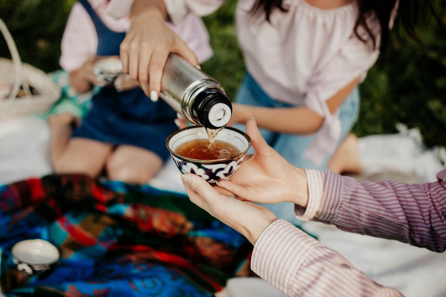 Eine Person gießt Tee aus einer Thermoskanne in eine gemusterte Tasse, die von einer anderen Person gehalten wird. Sie sitzen auf einer Picknickdecke im Freien, mit verschwommenen Figuren im Hintergrund.