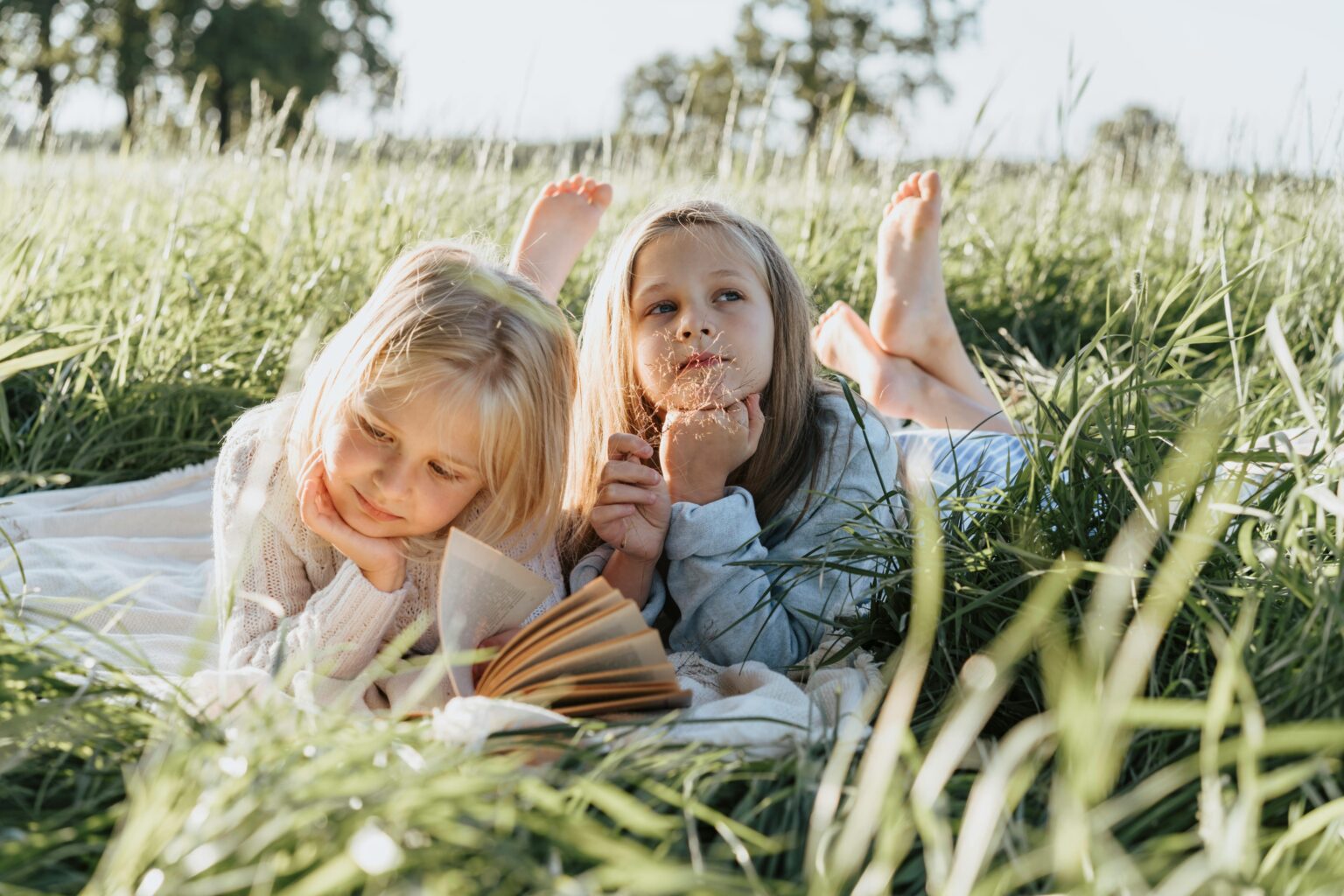 Zwei junge Mädchen liegen auf einer Decke im hohen Gras, die eine liest ein Buch, die andere hält spielerisch einen Löwenzahn. Das Sonnenlicht fällt sanft auf die beiden und schafft eine entspannte, sommerliche Atmosphäre.