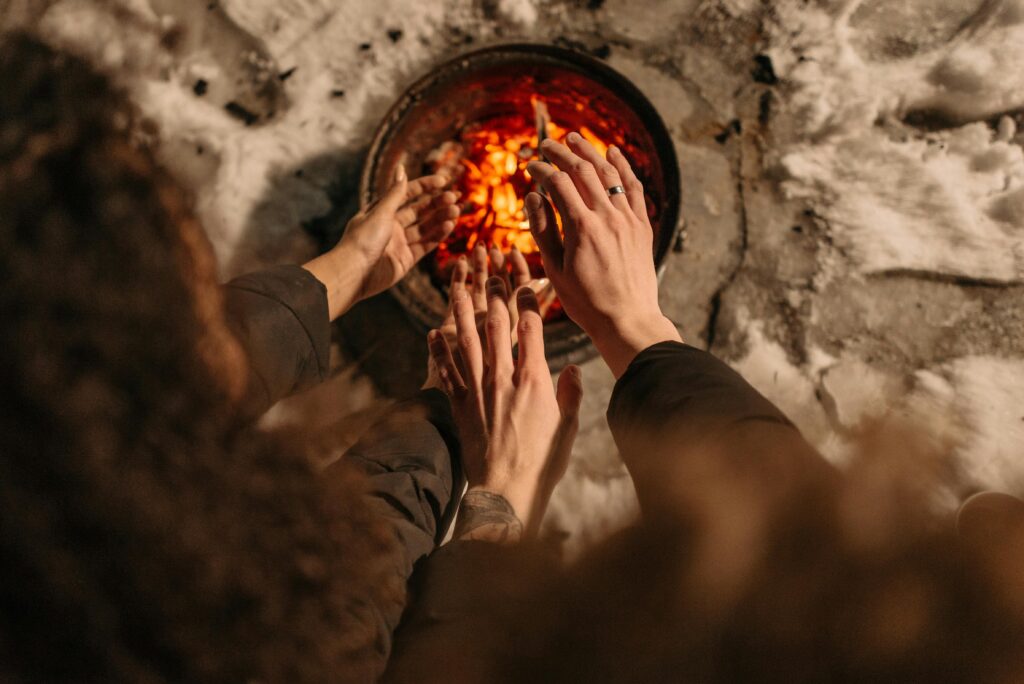 Mehrere Personen wärmen ihre Hände an einer glühenden Feuerstelle im Freien, umgeben von Schnee. Ihre Hände sind der Wärme entgegengestreckt, und die Szene fängt einen gemütlichen Moment in einer kalten Umgebung ein.