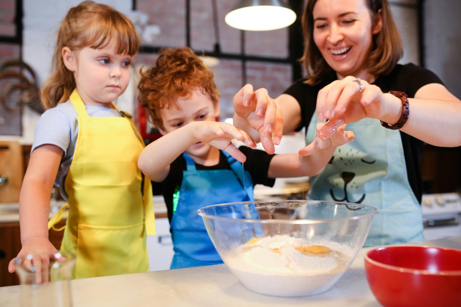 Eine Frau und zwei kleine Kinder schlagen in einer Küche Eier in eine durchsichtige, mit Mehl gefüllte Rührschüssel. Die Kinder tragen gelbe und blaue Schürzen, und die Frau lächelt, während sie ihnen beim gemeinsamen Kochen hilft.