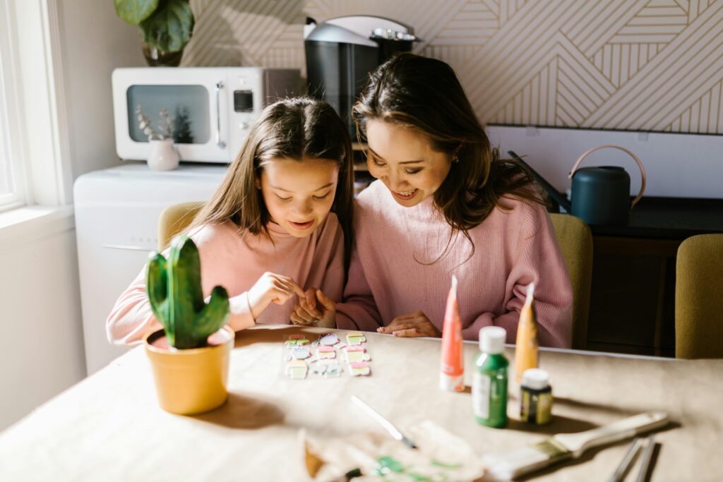 Eine Frau und ein Mädchen sitzen an einem Tisch und verzieren Kekse mit buntem Zuckerguss und lächeln dabei. Auf dem Tisch in einer gemütlichen, gut beleuchteten Küche stehen Malutensilien und ein getöpferter Kaktus.