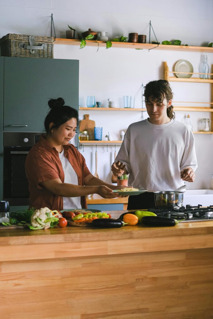 Zwei Personen bereiten gemeinsam in einer modernen Küche das Essen zu, mit frischem Gemüse auf der Holztheke, offenen Regalen mit Geschirr und grünen Schränken im Hintergrund.
