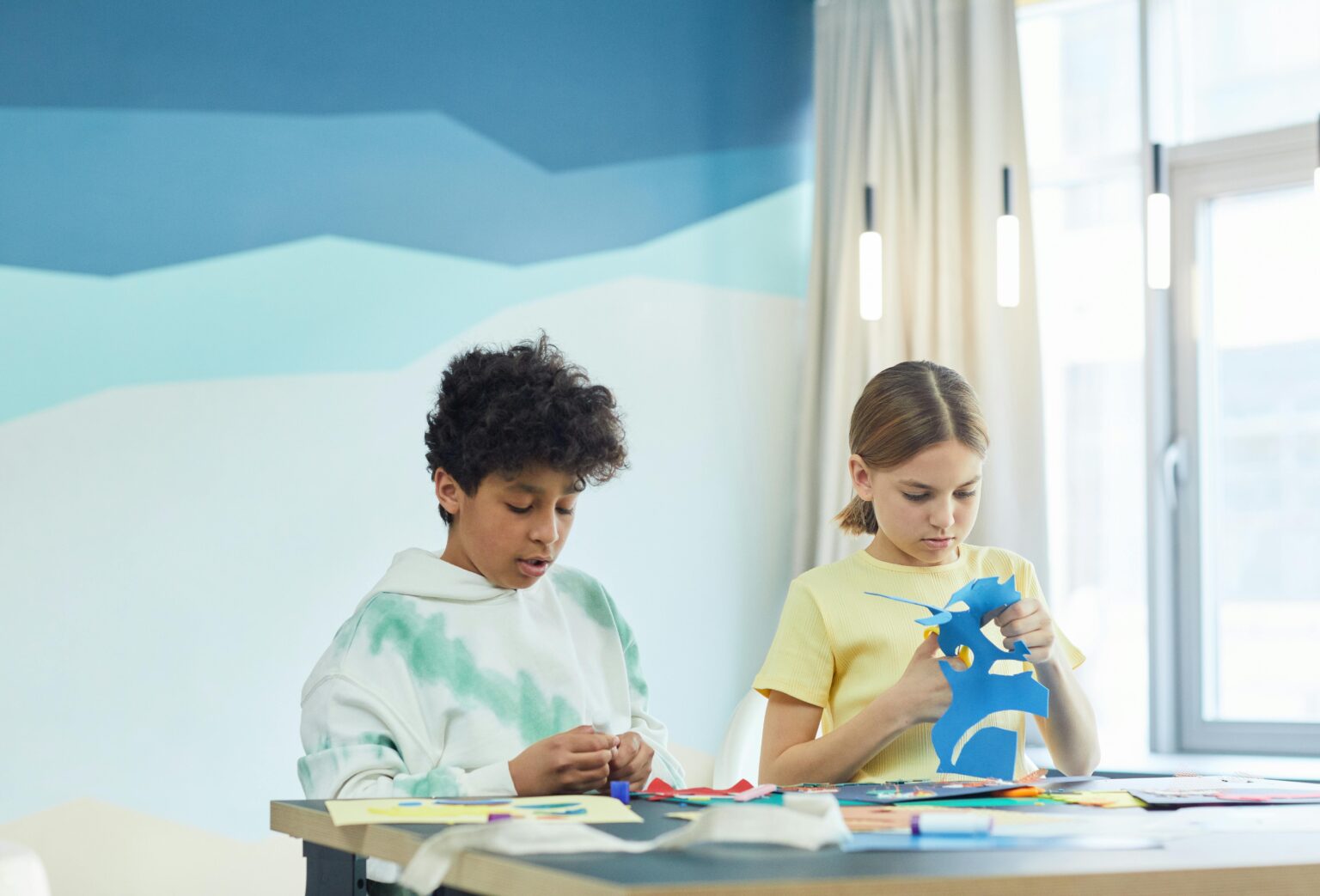 Zwei Kinder sitzen an einem Tisch und basteln. Ein Junge arbeitet mit farbigem Papier, während das Mädchen neben ihm mit einer Schere blaues Papier ausschneidet. In einem hell erleuchteten Raum sind sie auf ihre Projekte konzentriert.