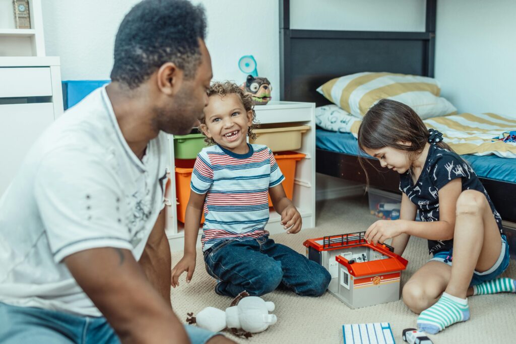 Ein Mann und zwei kleine Kinder sitzen in einem Schlafzimmer auf dem Boden, lächeln und spielen zusammen mit Spielzeug neben einem Bett mit einer gelb-weiß gestreiften Decke.
