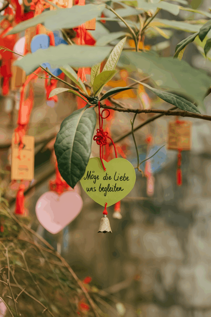 Ein grüner herzförmiger Anhänger mit deutschem Text, roter Schnur und einer kleinen Glocke hängt an einem Baumzweig, umgeben von anderen bunten Anhängern und roten Bändern.