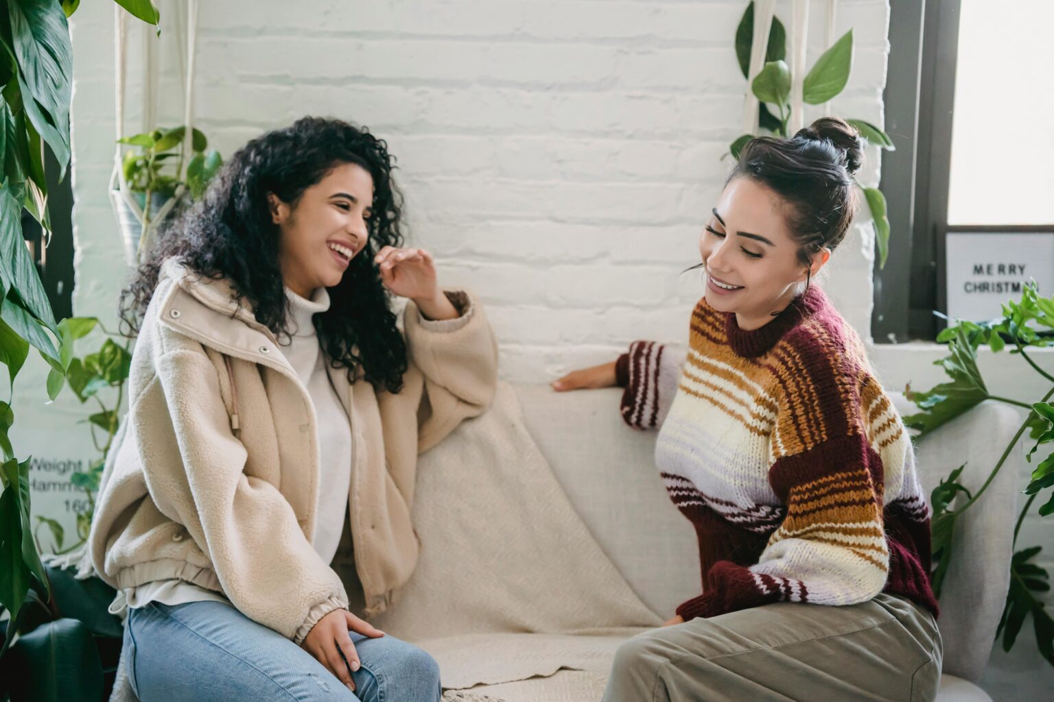 Zwei Frauen sitzen auf einer Couch, lächeln und reden miteinander, umgeben von grünen Zimmerpflanzen in einem hellen, gemütlichen Raum.