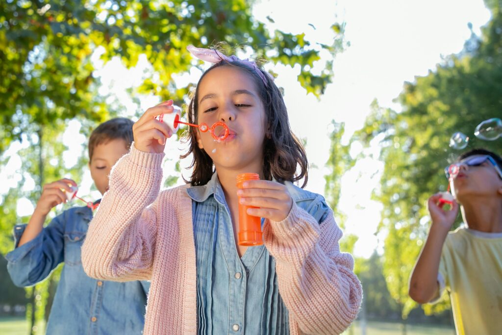 Drei Kinder sind im Freien und blasen Seifenblasen, während das Sonnenlicht durch die grünen Bäume im Hintergrund fällt. Im Mittelpunkt steht ein Mädchen in einem rosa Pullover, das Seifenblasenlösung in der Hand hält.