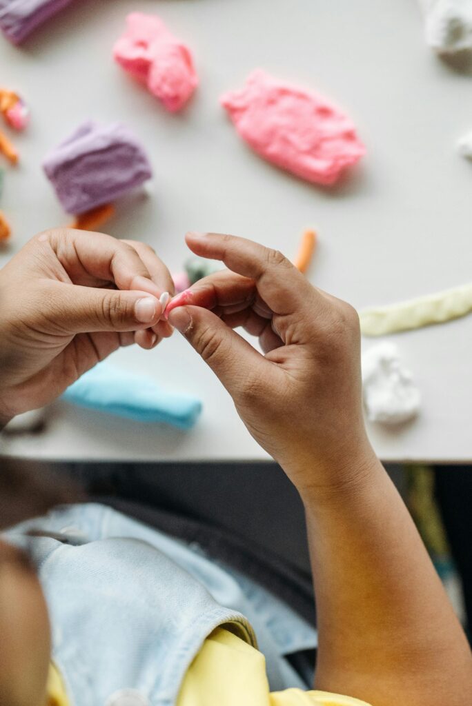 Die Hände eines Kindes formen bunte Knetmasse auf einem weißen Tisch, auf dem verschiedene Tonstücke verstreut sind.