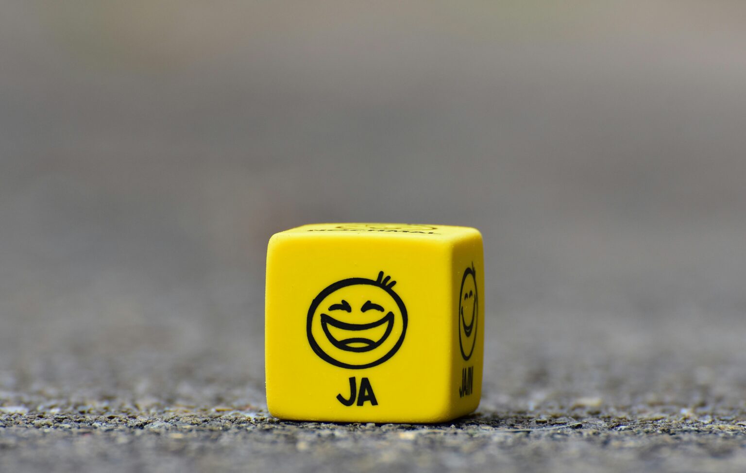 Ein gelber Würfel mit einem lächelnden Gesichtsemoji und dem Wort JA sitzt auf einer grauen, leicht strukturierten Oberfläche.