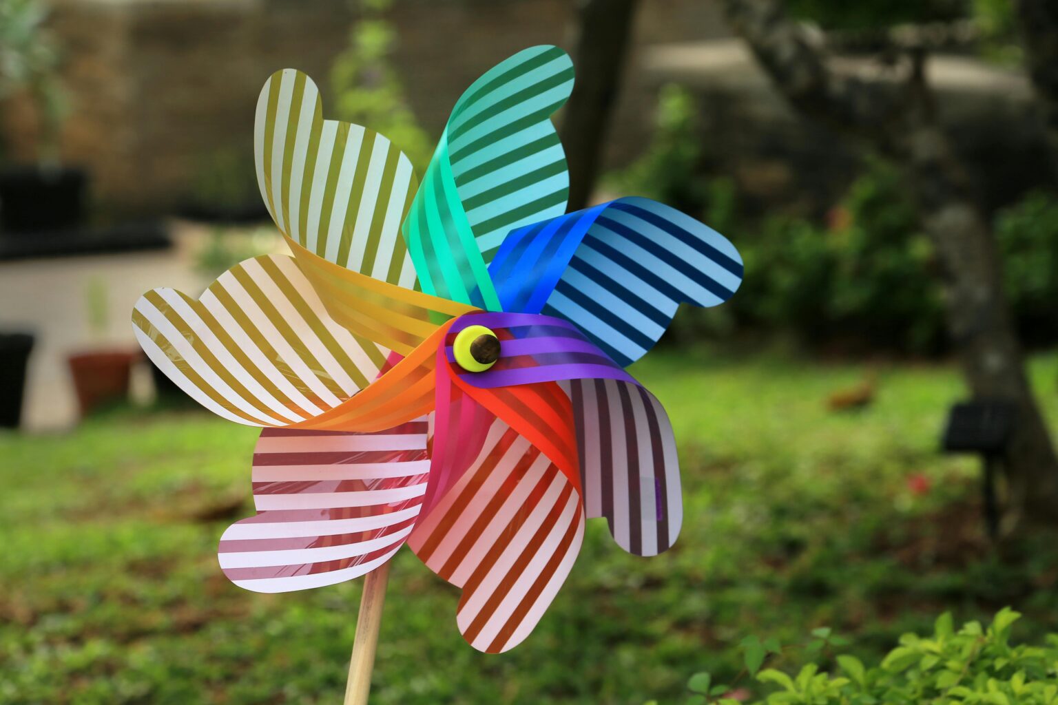 Ein bunt gestreiftes Windrad dreht sich auf einem Stock in einem Garten, mit grünem Gras und verschwommenen Bäumen im Hintergrund.