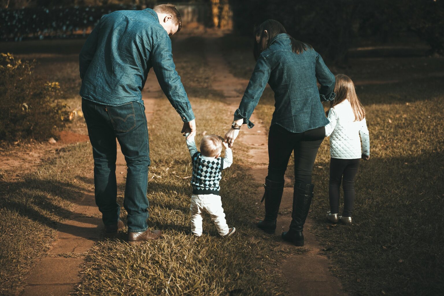 Eine vierköpfige Familie, von hinten gesehen, geht einen Weg entlang. Zwei Erwachsene halten ein Kleinkind an der Hand, während ein älteres Kind neben ihnen läuft. Alle tragen Freizeitkleidung.