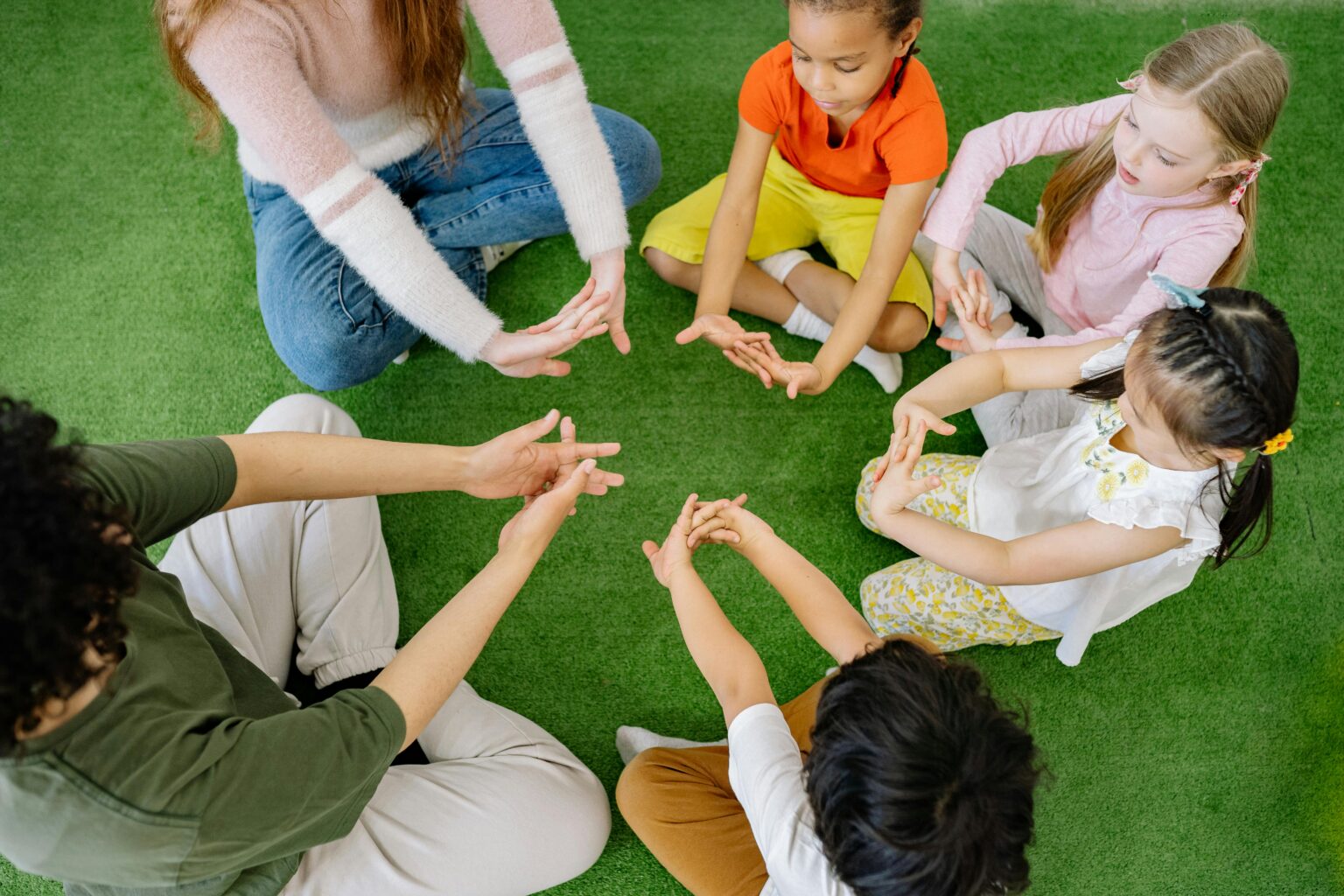 Fünf kleine Kinder und ein Erwachsener sitzen im Kreis auf einem grünen Teppich, berühren sich an den Händen und spielen gemeinsam ein Gruppenspiel.
