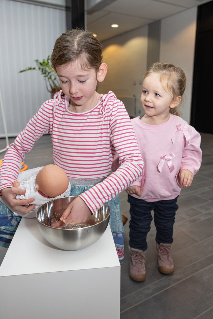Zwei junge Mädchen stehen in einem Haus; die eine schlägt ein Ei in eine Metallschüssel auf, während die andere lächelnd zusieht. Beide tragen legere Kleidung und stehen auf einem grau gekachelten Boden.
