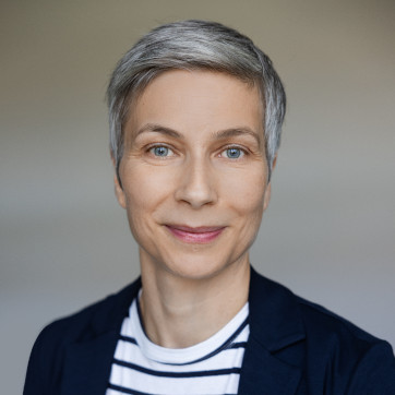 Eine Person mit kurzen grauen Haaren, die einen dunklen Blazer über einem weiß-schwarz gestreiften Hemd trägt, lächelt sanft vor einem neutralen Hintergrund.