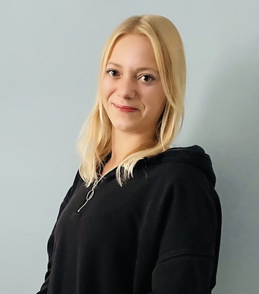 Eine Frau mit glattem blondem Haar, die einen schwarzen Kapuzenpulli trägt, steht vor einer schlichten hellblauen Wand und lächelt sanft in die Kamera.