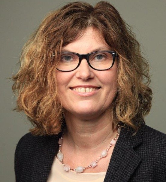 Eine Frau mit gewelltem, schulterlangem hellbraunem Haar, die eine Brille, einen dunklen Blazer, ein helles Oberteil und eine Perlenkette trägt, lächelt vor einem schlichten Hintergrund.