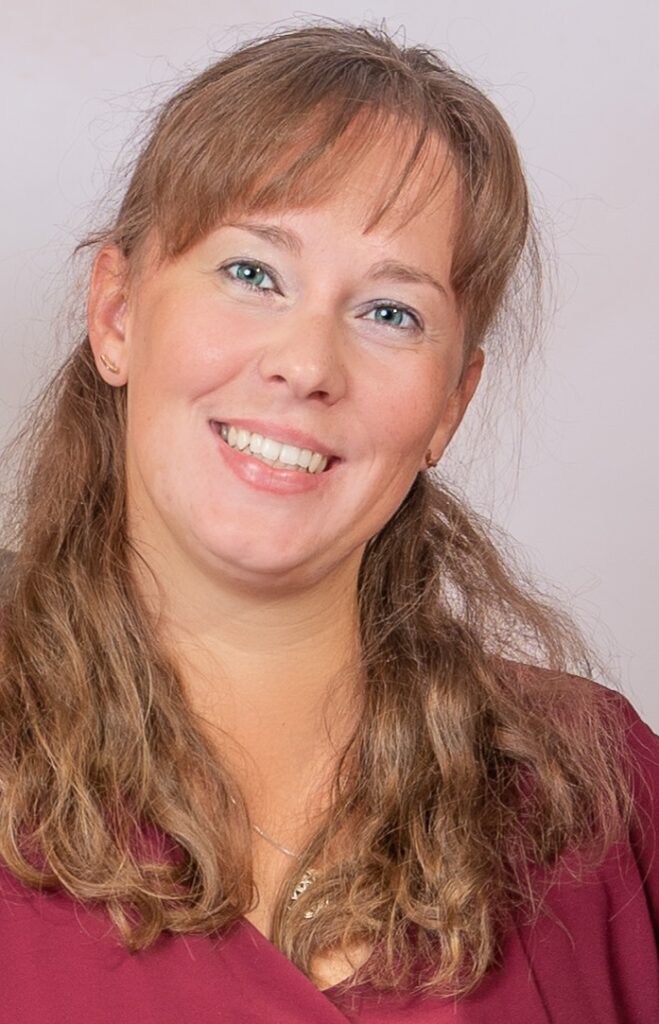 Eine Frau mit langem hellbraunem Haar und Pony lächelt in die Kamera. Sie trägt ein kastanienbraunes Oberteil und kleine Ohrringe und steht vor einem hellen Hintergrund.