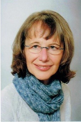 Eine Frau mit schulterlangem hellbraunem Haar, Brille und einem blau gemusterten Schal lächelt in die Kamera vor einem hellen Hintergrund.