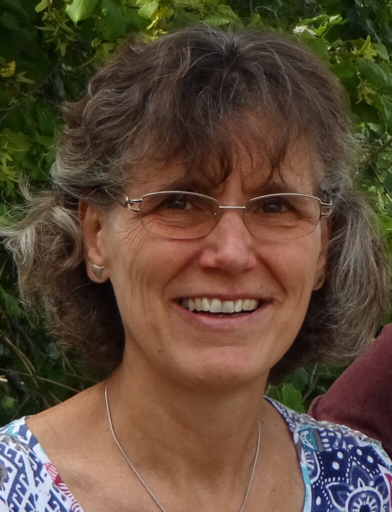 Eine Frau mit lockigem braunem Haar, Brille und gemustertem Hemd lächelt im Freien vor grünem Laub.