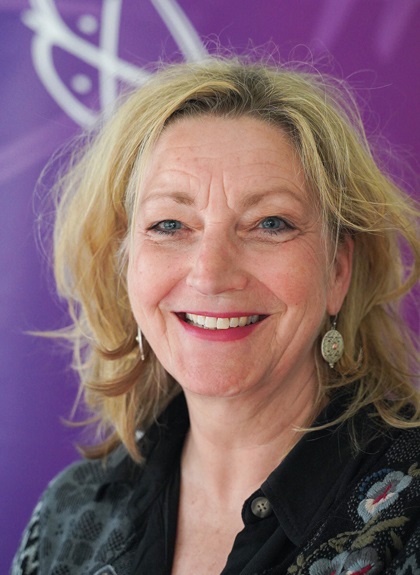 Eine Frau mit blondem Haar lächelt in die Kamera. Sie trägt Ohrringe und ein schwarz gemustertes Oberteil vor einem lila Hintergrund mit einem weißen abstrakten Muster.