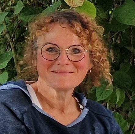 Eine lächelnde Frau mit lockigem Haar und runder Brille sitzt im Freien vor grünen Blattpflanzen und trägt einen marineblauen Kapuzenpullover.