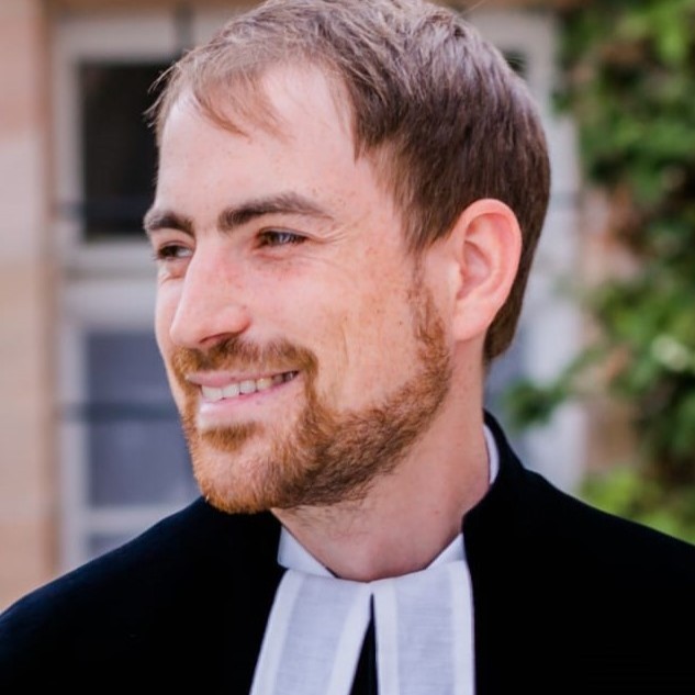 Ein Mann mit kurzen braunen Haaren und einem Bart, der ein schwarzes Gewand mit weißem Kragen trägt, lächelt und blickt zur Seite ins Freie.