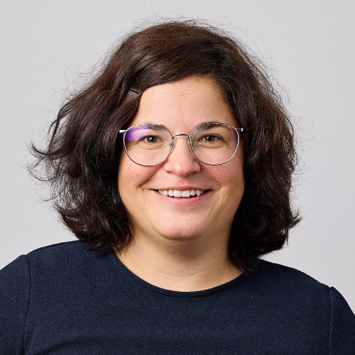 Eine Person mit schulterlangem dunklem Haar und Brille, lächelnd, mit dunklem Oberteil, vor einem schlichten hellgrauen Hintergrund.