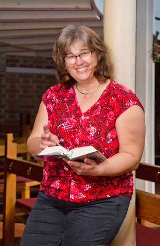 Eine lächelnde Frau mit Brille und rot gemusterter Bluse hält ein aufgeschlagenes Buch in einem warm beleuchteten Innenbereich mit Holzstühlen im Hintergrund.