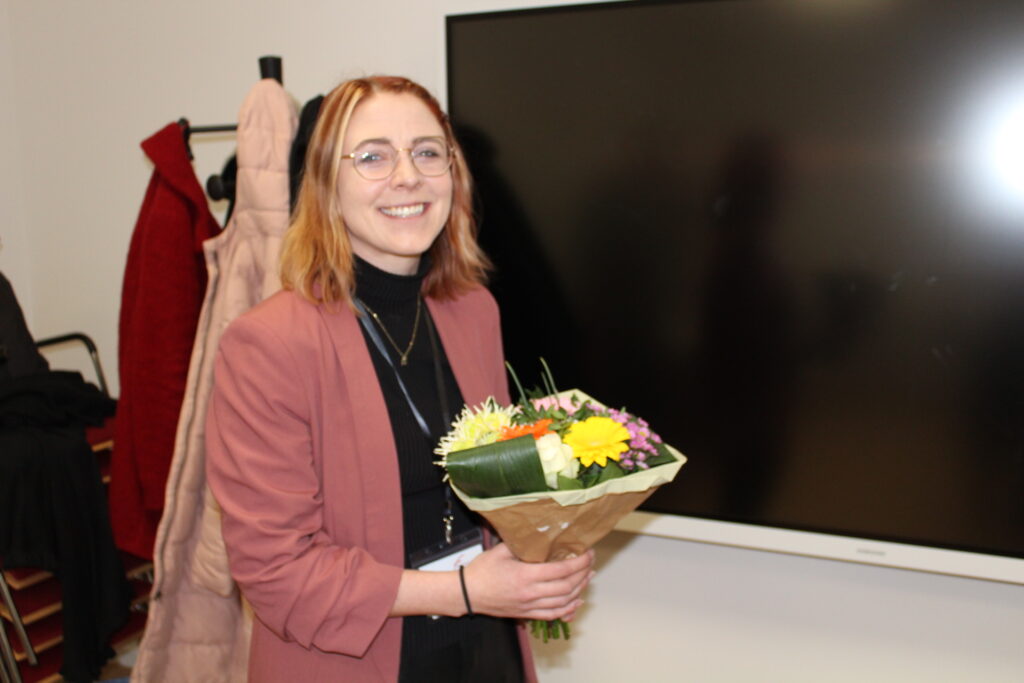 Eine lächelnde Person mit Brille und hellbraunem Haar hält einen Blumenstrauß in der Hand und steht in einem Innenraum neben einem großen Bildschirm und einer Garderobe.