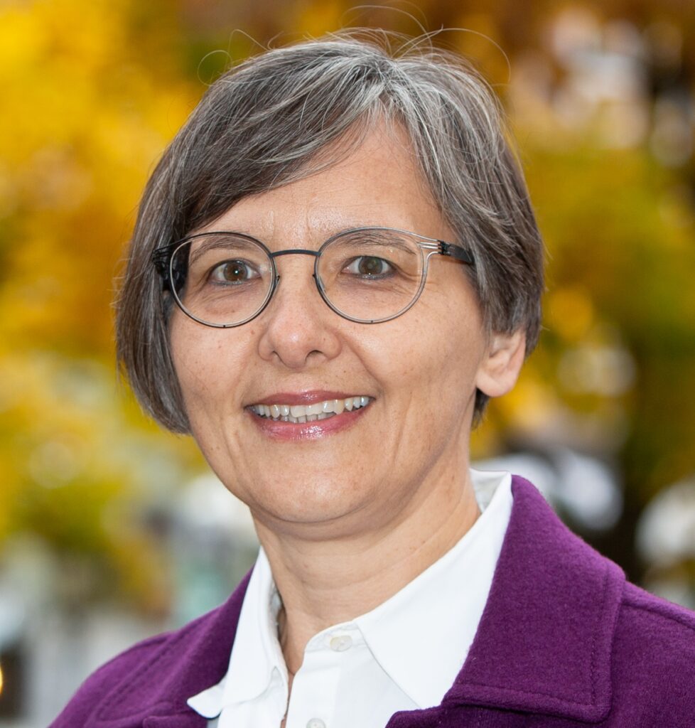 Lächelnde Frau mit kurzen grauen Haaren und Brille, die ein weißes Hemd und einen lila Mantel trägt, steht im Freien vor einem verschwommenen gelben und grünen Hintergrund.