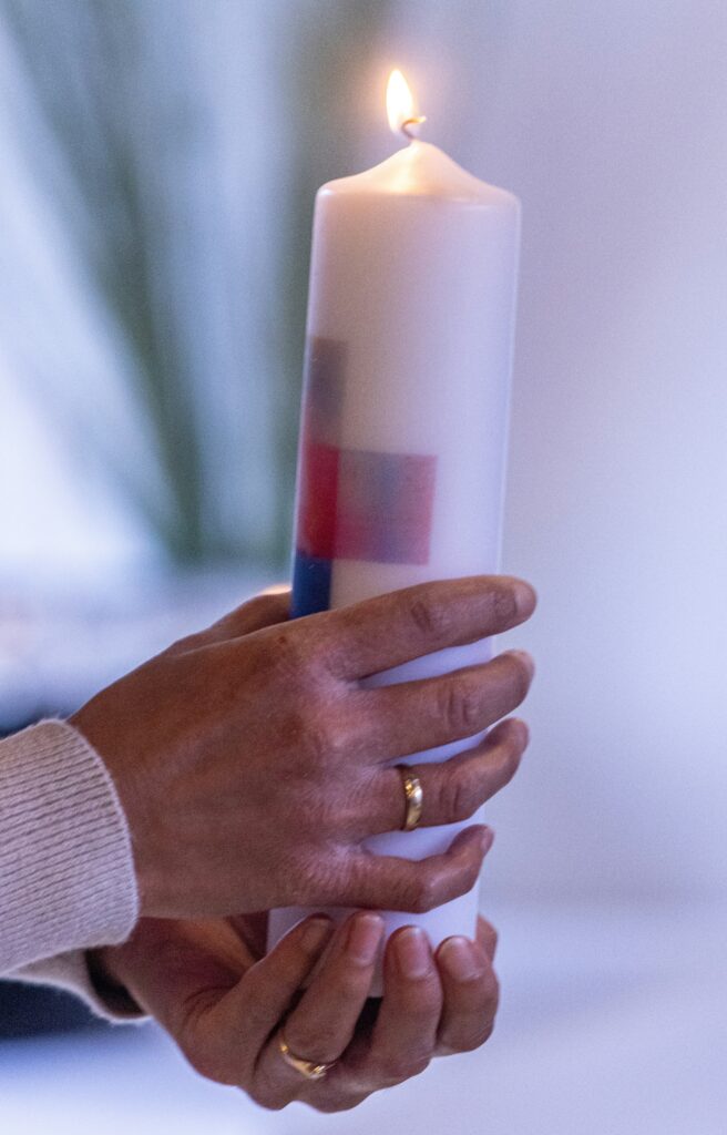Eine Person mit einem goldenen Ring hält eine brennende weiße Kerze, die mit roten und blauen Quadraten verziert ist, vor einem sanft verschwommenen Hintergrund.
