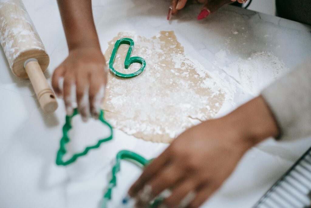 Zwei Personen formen den Teig mit grünen Ausstechern auf einer bemehlten Fläche, ein Nudelholz liegt daneben, und bereiten gemeinsam festliche Plätzchen zu.