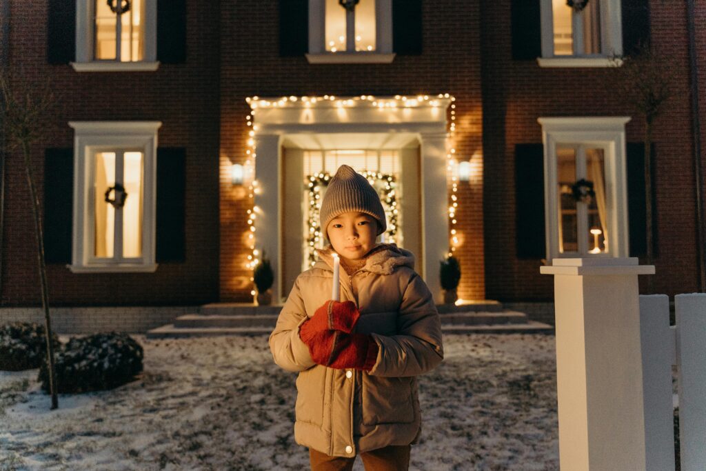 Ein Kind in Wintermantel und Mütze hält eine brennende Kerze vor einem mit Lichtern und Kränzen geschmückten Haus, das in der Dämmerung auf einem verschneiten Hof steht.