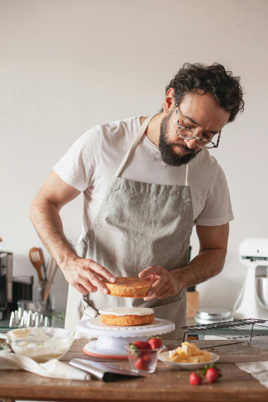 Ein Mann mit Brille und Schürze setzt in einer Küche eine Torte zusammen, indem er vorsichtig eine Schicht auf die andere legt.