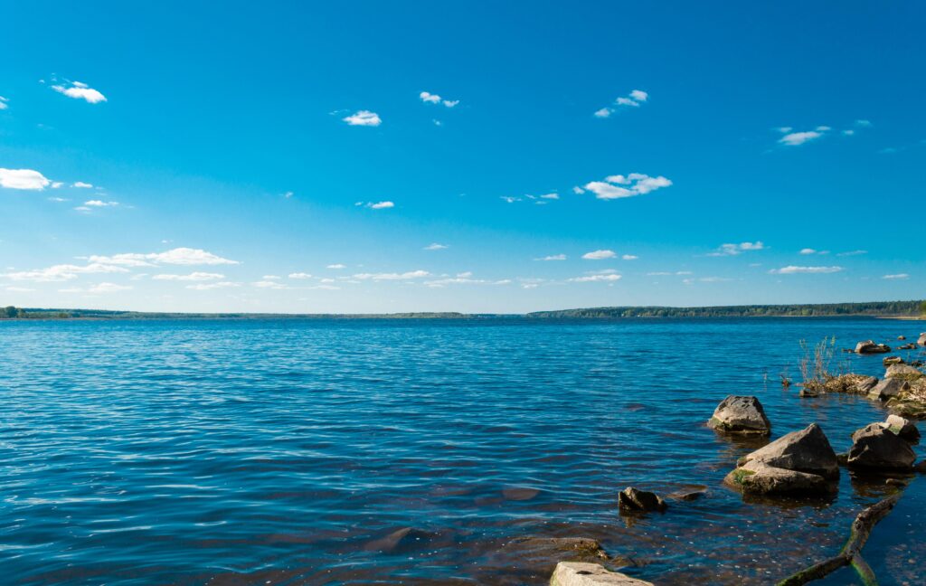 Ein ruhiger See mit klarem, blauem Wasser, verstreute Felsen im Vordergrund und ein entferntes, von Bäumen gesäumtes Ufer unter einem strahlend blauen Himmel mit ein paar kleinen Wolken.