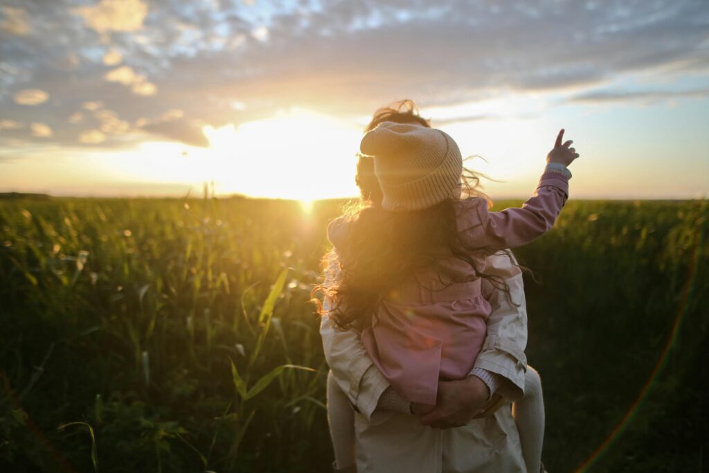 Ein Erwachsener trägt ein Kind auf einem Feld bei Sonnenuntergang. Das Kind, das einen Hut trägt, zeigt in den Himmel, während die Sonne hinter ihnen untergeht.