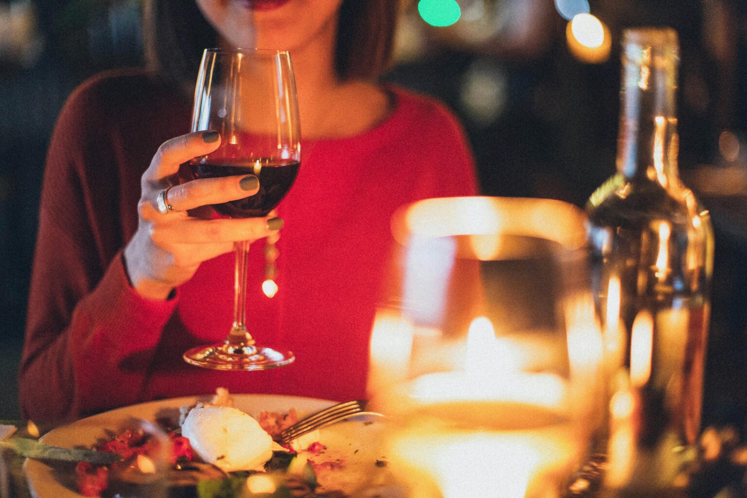 Eine Frau in einem roten Oberteil hält ein Glas Rotwein an einem mit Kerzen beleuchteten Tisch mit Speisen und einer Weinflasche, wodurch eine warme, intime Atmosphäre entsteht.