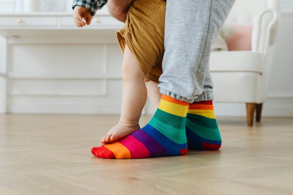 Ein Erwachsener, der Socken mit Regenbogenstreifen trägt, steht auf einem Holzboden, während ein kleines Kind auf seinen Füßen steht und sich an den Beinen festhält.