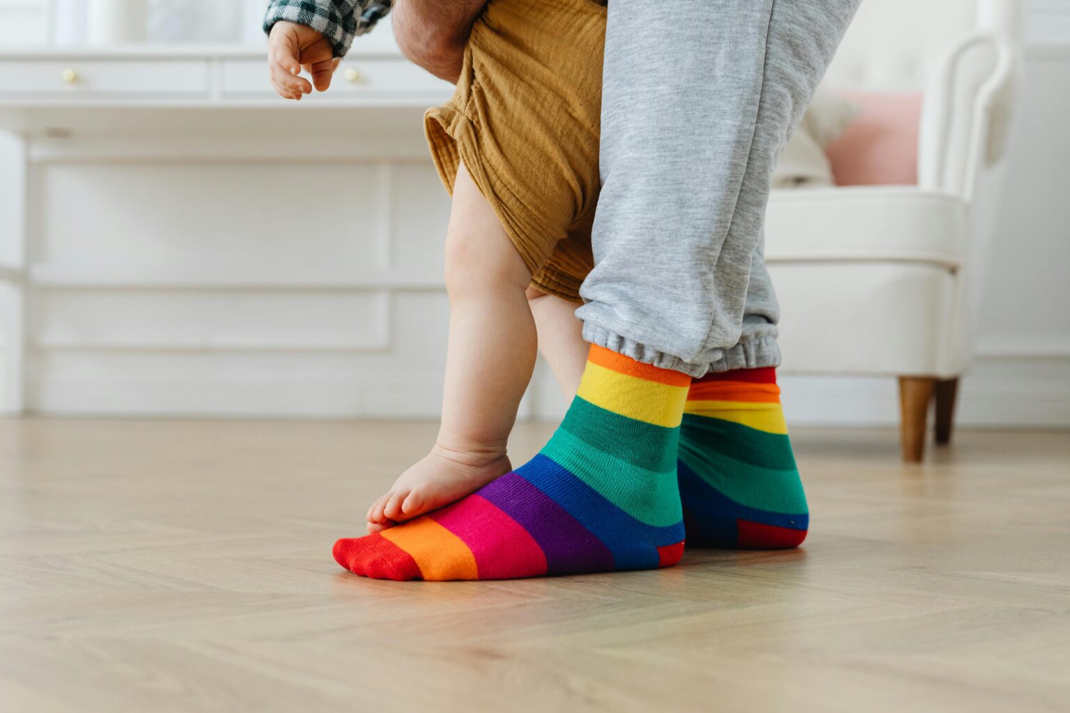 Ein Erwachsener, der Socken mit Regenbogenstreifen trägt, steht auf einem Holzboden, während ein kleines Kind auf seinen Füßen steht und sich an den Beinen festhält.
